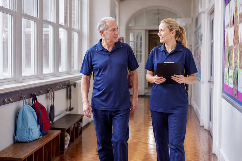 A school caretaker and a cleaning supervisor in matching uniforms inspecting a bright primary school corridor, one holding a clipboard with a cleaning schedule, natural daylight through tall windows