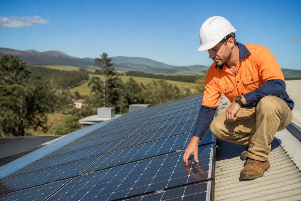 Certified solar installer in high-visibility work uniform examining photovoltaic panels on residential rooftop, blue sky and rural landscape visible in background