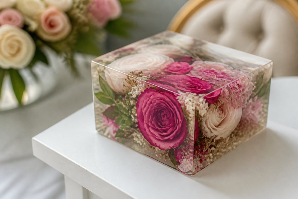 Close-up of a preserved wedding bouquet displayed in a custom resin keepsake on a white table, showing vibrant pink and white flowers with intricate petal detail and clear resin casting