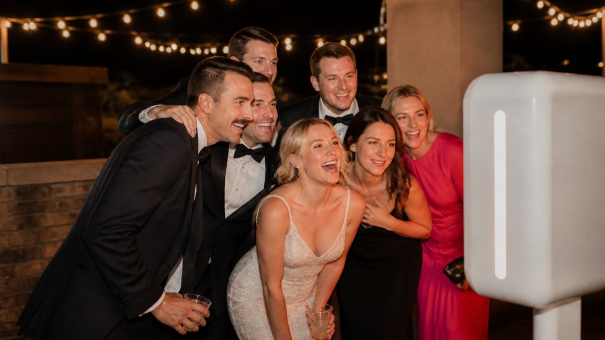 Wedding guests in formal attire laughing and posing together inside an open-air photo booth at an evening reception, warm string lights in the background