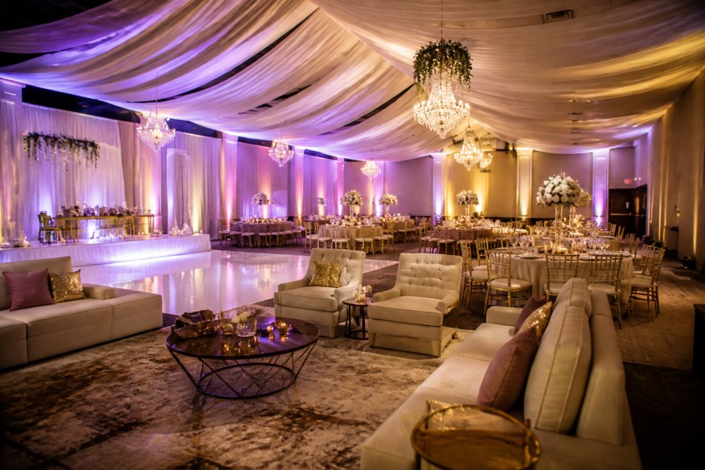 An elegantly decorated indoor wedding reception space with draped ceiling fabric, glowing up-lights in soft purple and gold, lounge furniture arranged near a dance floor, and round tables set for a formal dinner under warm ambient lighting