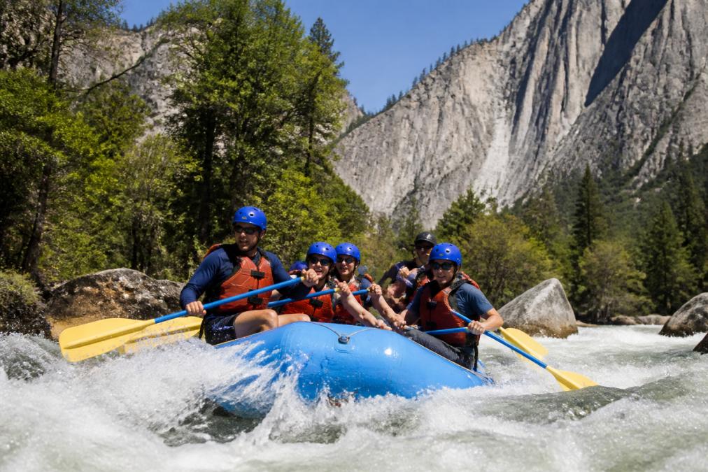 A guided raft navigating Class III rapids on the Merced River with granite cliffs visible in the background, paddlers in helmets and life vests working as a team