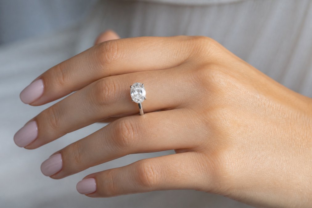 Close-up of a hand with slender fingers wearing an elegant oval-cut diamond engagement ring, soft natural lighting, neutral background, jewellery photography style