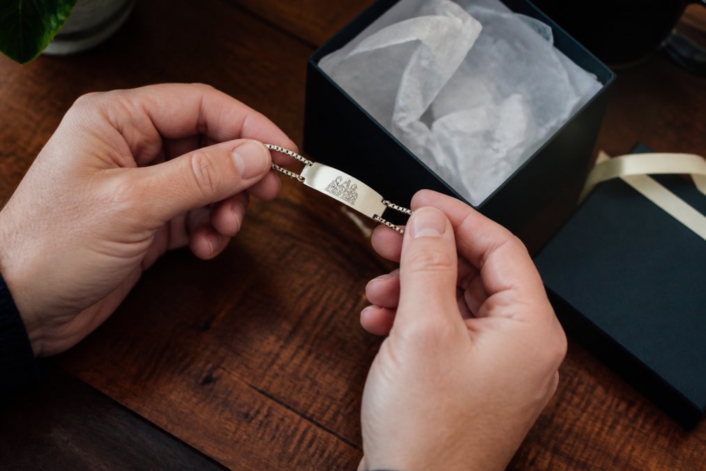 Close-up of hands holding an engraved stainless steel bracelet beside a small gift box with tissue paper, soft warm lighting on a wooden table