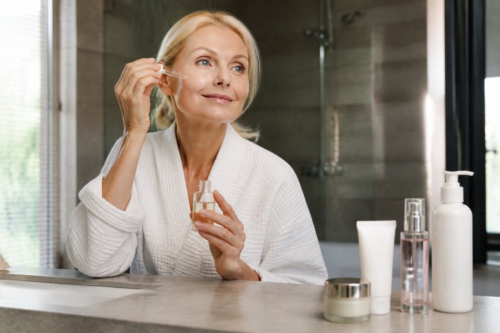 A woman in her 50s with glowing skin applying serum in a bright bathroom, surrounded by skincare products, looking confident and relaxed