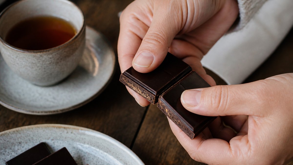 Close-up of hands breaking open an artisanal chocolate bar, revealing rich interior texture and snap, with a cup of tea nearby on a wooden surface