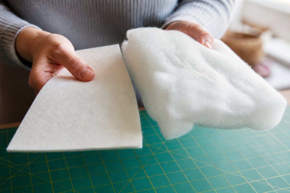 Close-up of a quilter's hands holding two batting samples in natural window light, one showing the flat matted texture of cotton and the other the fluffy resilient loft of polyester