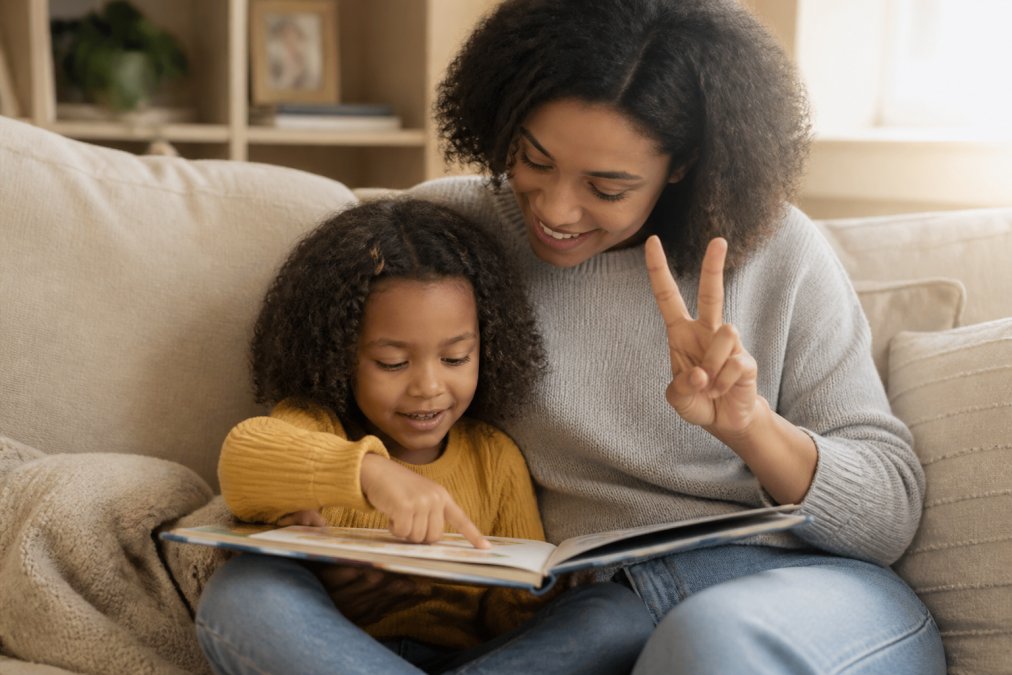 A parent and child sitting together on a cozy couch in warm afternoon light, holding an open picture book, the child pointing at a word while the parent smiles and holds up three fingers