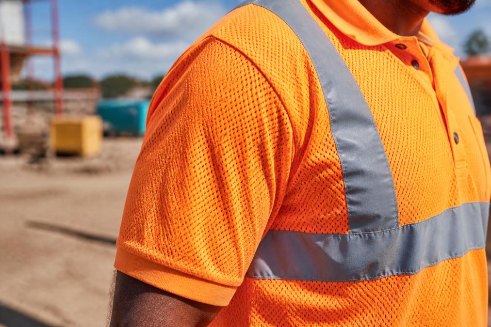 A close-up of a worker on a construction site wearing an orange hi vis short sleeve polo with reflective tape, showing the mesh fabric texture in natural daylight