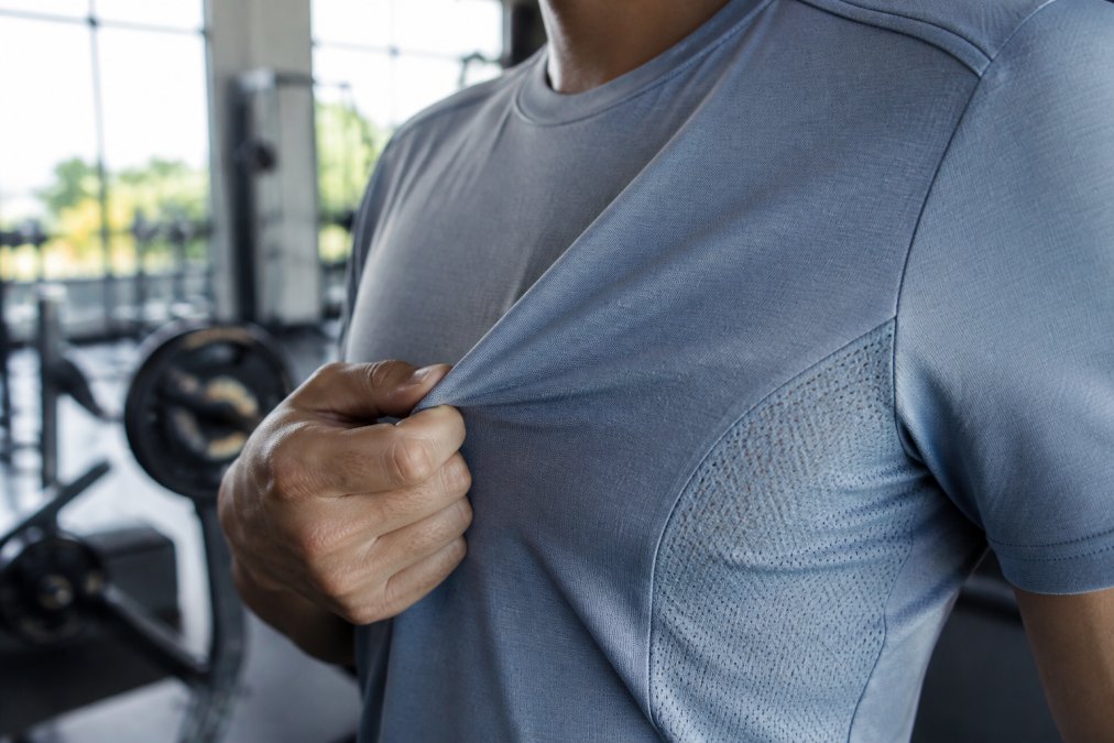 Close-up of a person's hand feeling the fabric of a lightweight athletic shirt, with mesh panels visible, in a bright gym setting