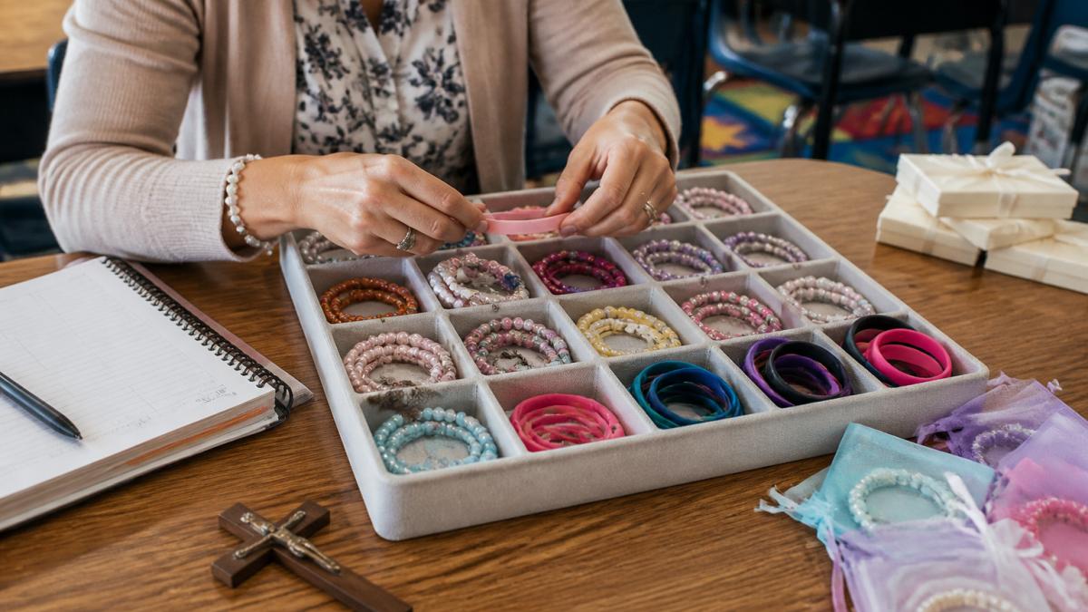 A teacher's hands sorting a tray of colorful silicone wristbands and beaded bracelets on a classroom table, with a lesson planner and a small cross visible nearby