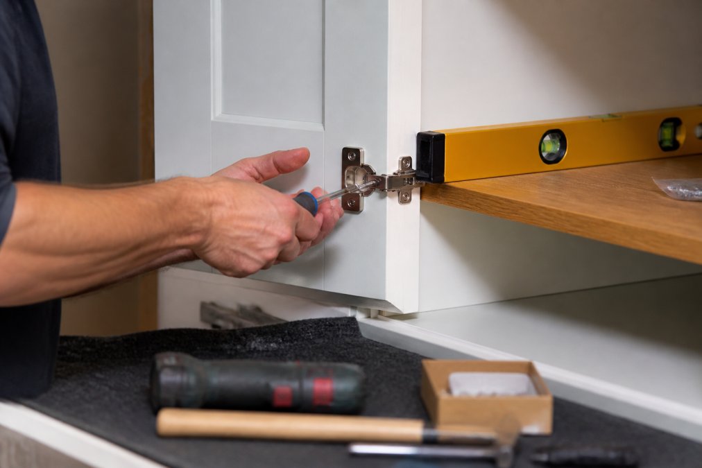 A joiner using a screwdriver to adjust cabinet hinges with a spirit level resting on the shelf edge.