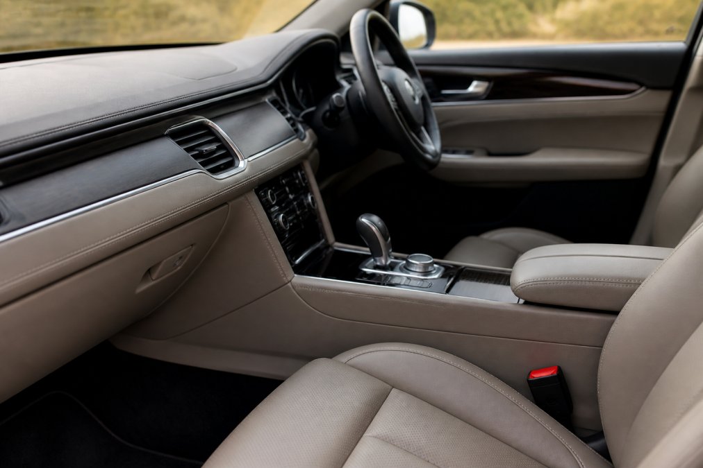 Close-up of a car interior dashboard and seats showing a cohesive neutral color palette with leather upholstery, stitching details, and metallic trim under natural cabin lighting