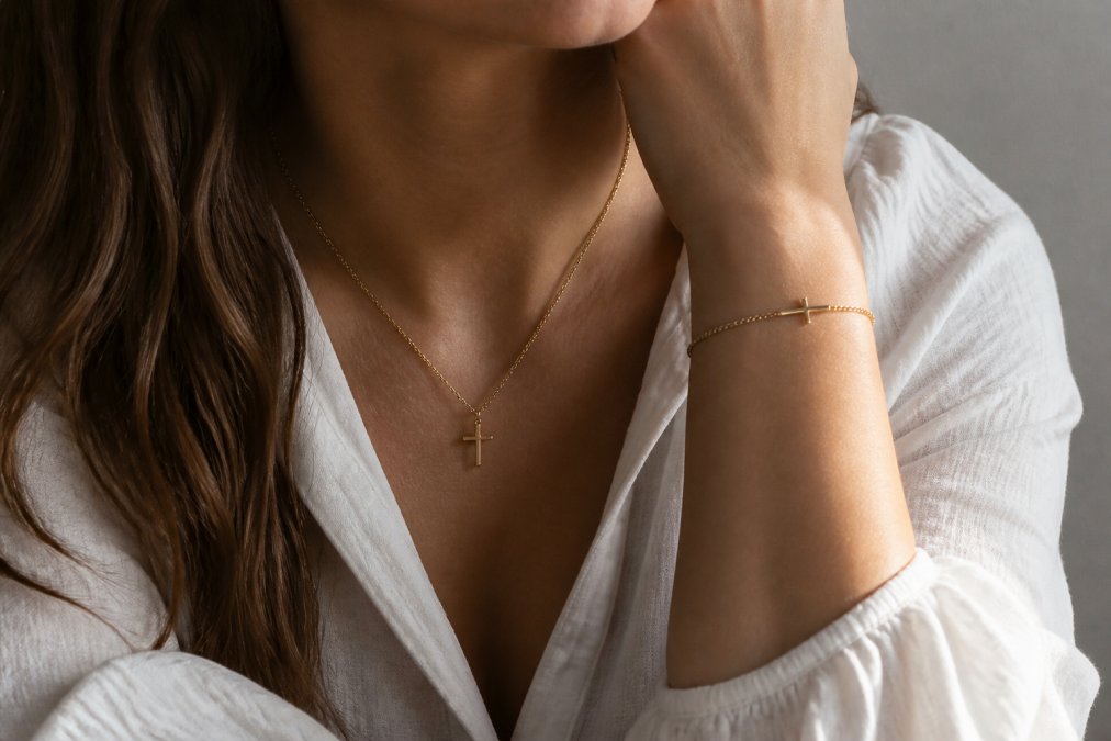 Close-up of a woman's wrist and neck wearing a delicate gold cross necklace and a simple bracelet, soft natural light, neutral background