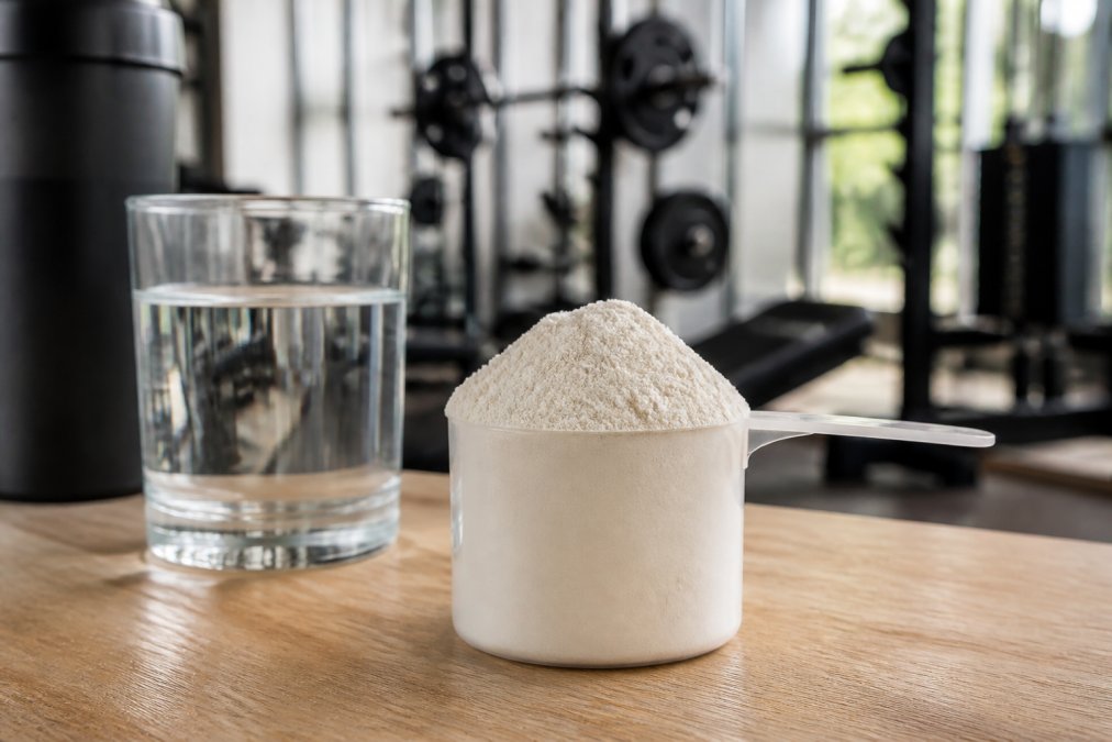 Close-up of a clean white protein powder scoop resting on a wooden surface next to a glass of water, with a blurred gym background in soft focus behind it