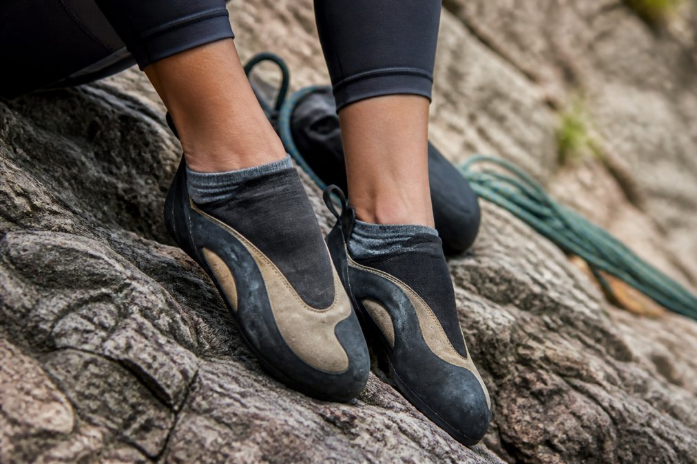 Close-up of a climber's feet wearing thin, snug climbing socks inside climbing shoes, resting on a textured rock surface at an outdoor crag
