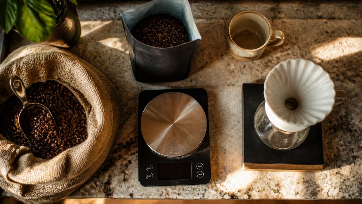 Overhead shot of a kitchen counter with a bag of whole bean coffee, a burlap sack, a digital scale, a pour-over dripper, and a coffee mug, warm morning light