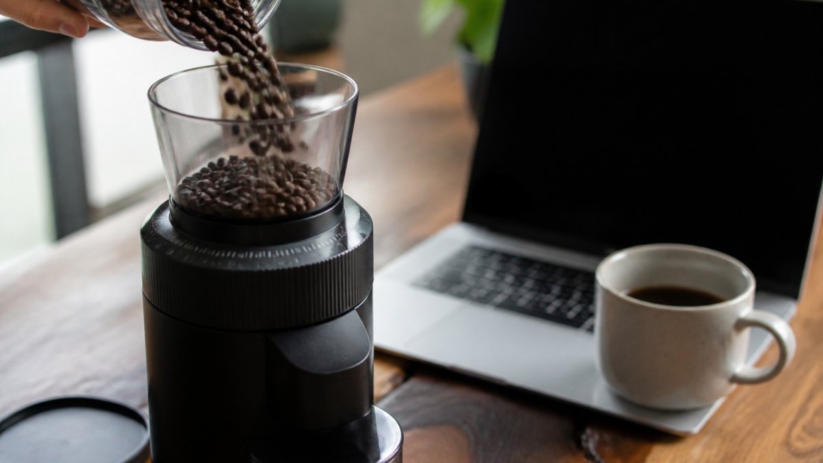 Whole coffee beans pouring into a burr grinder on a desk with a laptop and ceramic mug