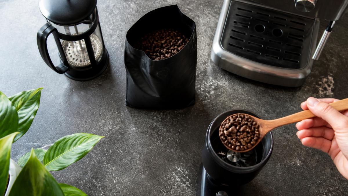 Overhead shot of a home kitchen bench with a French press, espresso machine and a bag of whole coffee beans, with a hand scooping beans into a grinder