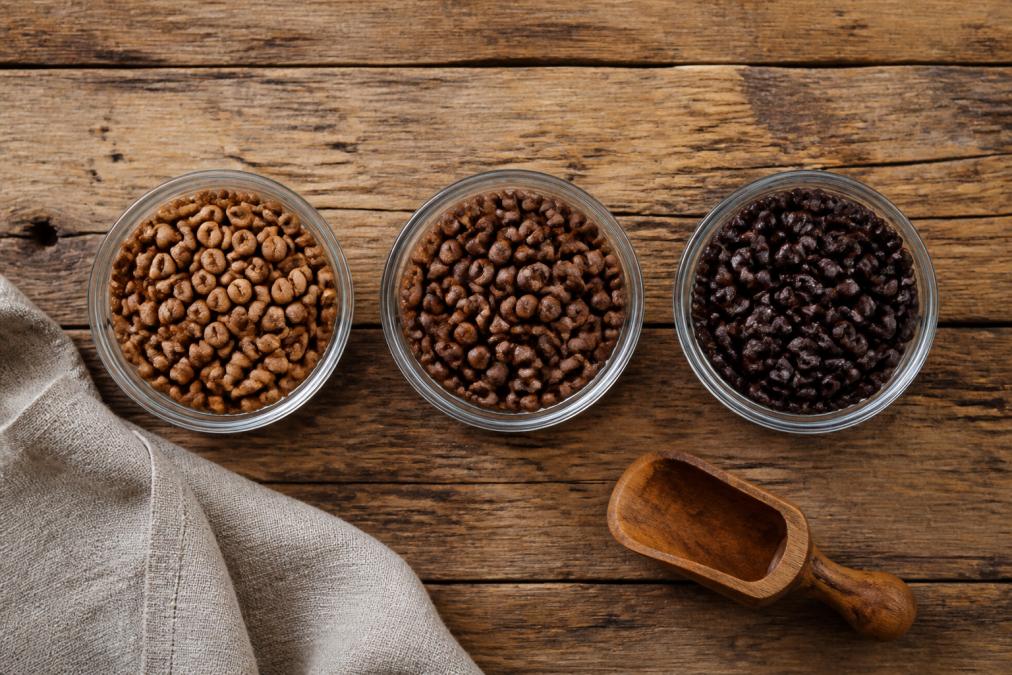 Overhead flat lay of three small glass bowls of whole coffee beans in light, medium, and dark roast shades arranged in a row on a rustic wooden table, with a linen napkin and a wooden scoop nearby