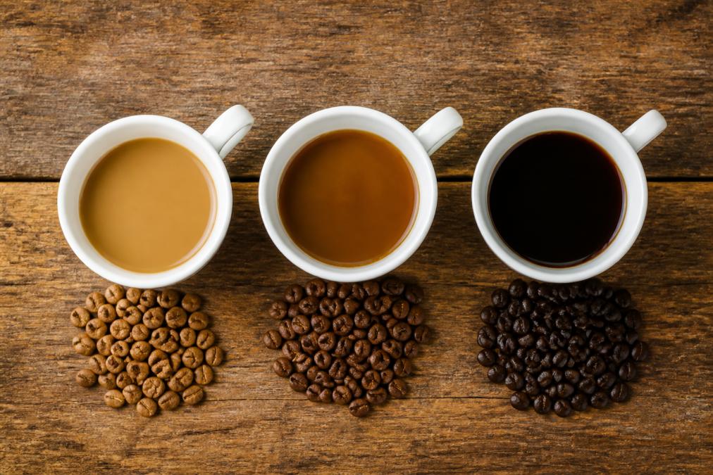 Three white ceramic cups arranged in a line from left to right, each containing brewed coffee showing distinct color progression from light tan to dark chocolate brown, with scattered whole coffee beans in their natural states beside each cup on a wooden surface, shot from directly above with natural window lighting
