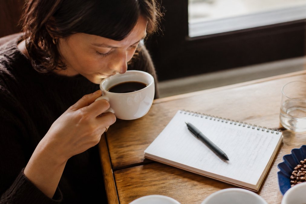 Person holding a white ceramic coffee cup near their nose with eyes closed, demonstrating aroma evaluation technique, with a handwritten tasting notepad and pen visible on a wooden table beside the cup, warm natural lighting from a nearby window