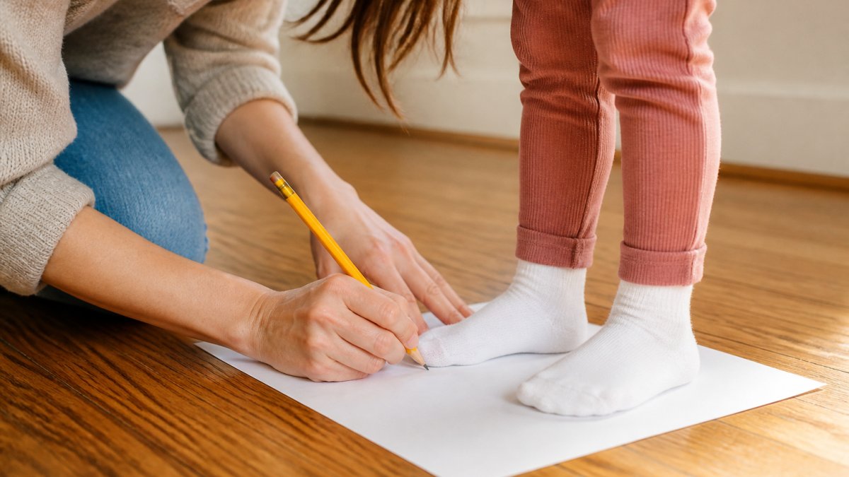 Parent kneeling on a hardwood floor marking the heel and toe of a toddler standing on paper to measure foot size