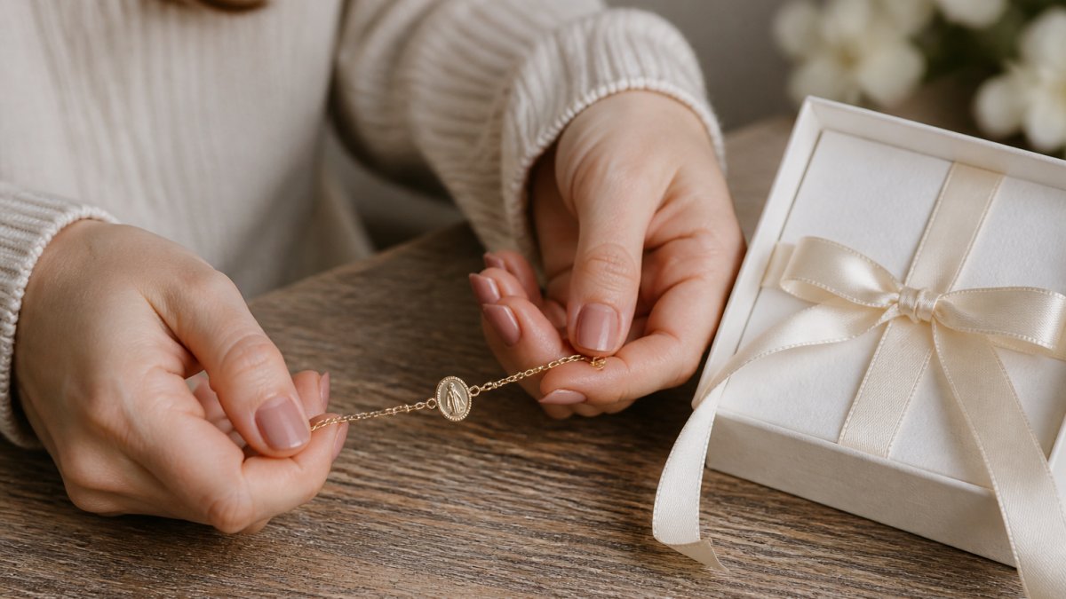 Close-up of a young woman's hands holding a delicate gold Miraculous Medal bracelet next to a white gift box on a wooden table, soft natural light