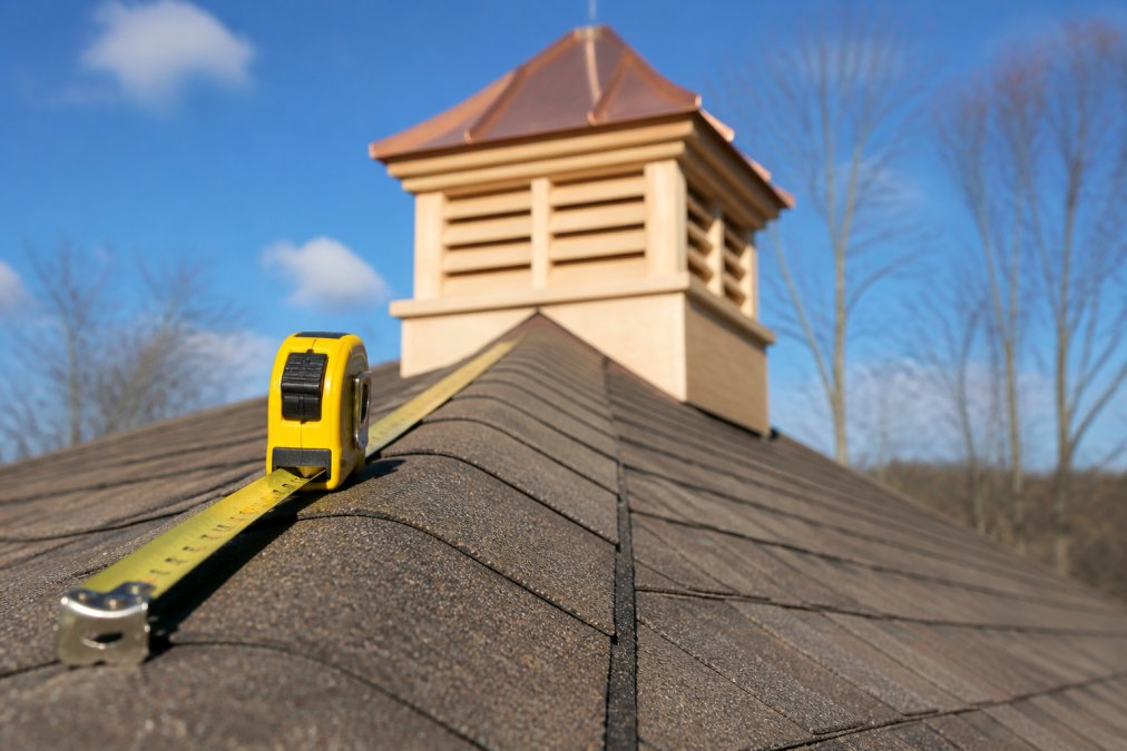 A close-up of a measuring tape extended along the ridge line of a residential roof, with a wooden cupola partially visible in the background under clear skies