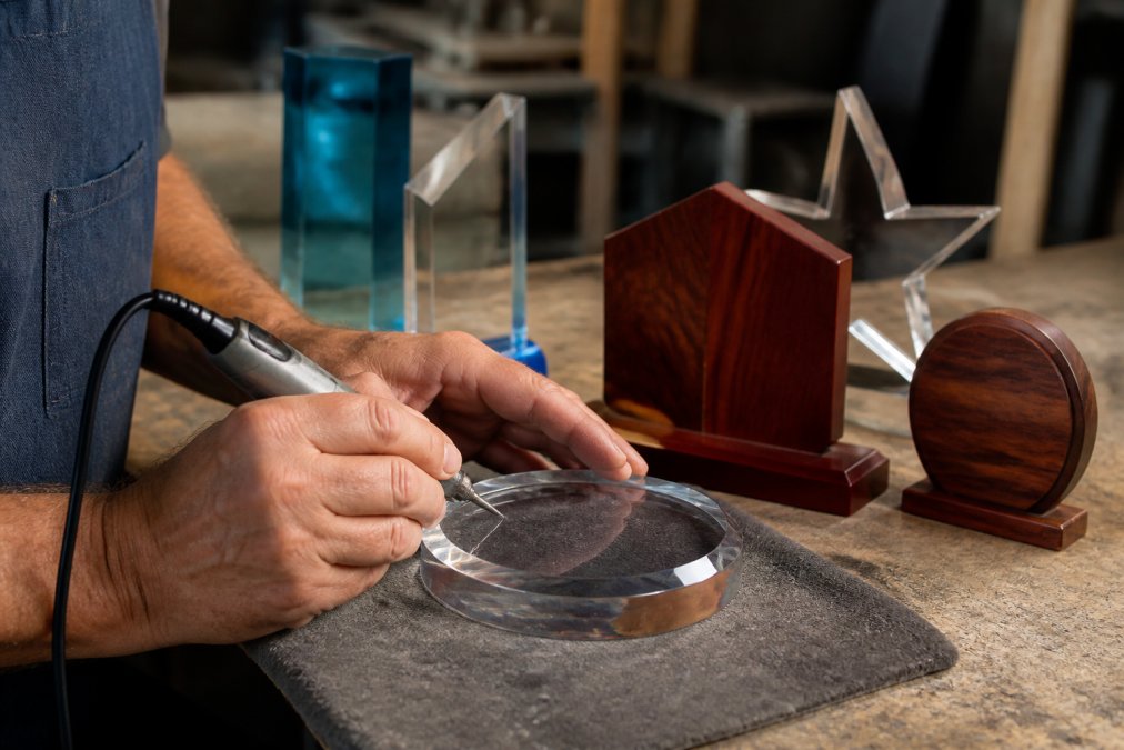 Craftsman engraving a crystal plaque for custom awards in a workshop with wood and acrylic materials nearby