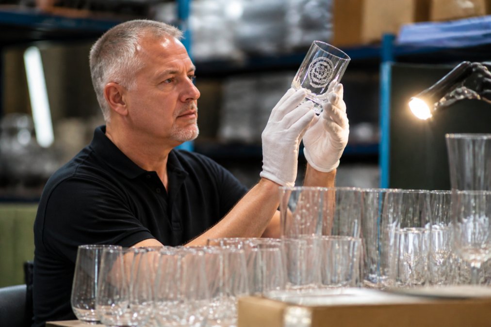 Quality control technician carefully inspecting custom-branded glassware under bright lighting, examining etching clarity, surface finish, and branding precision on multiple pieces