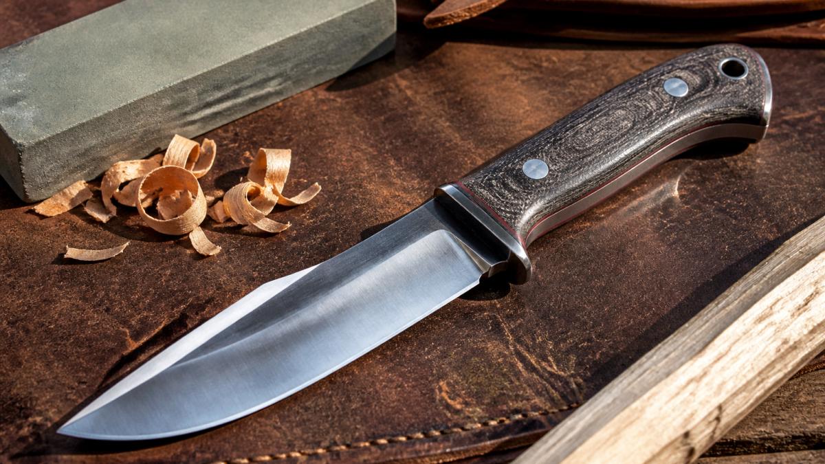 Custom hunting knife with a flat grind resting on weathered leather beside a sharpening stone and wood shavings