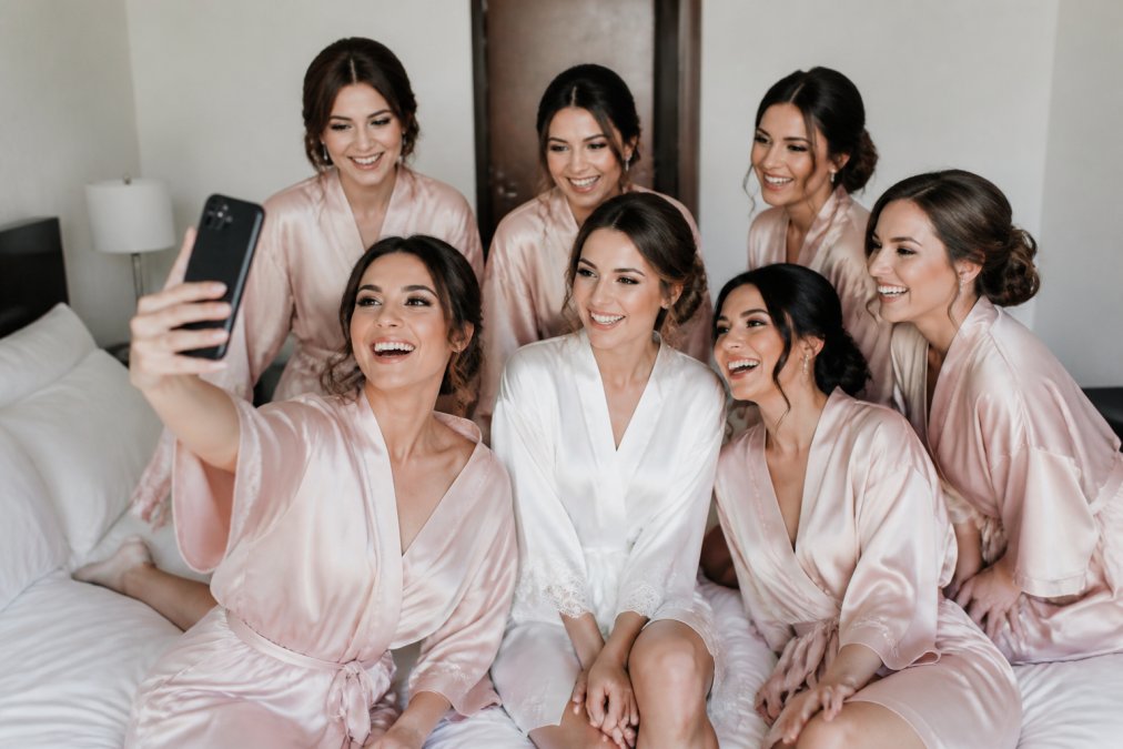 Bridesmaids in matching custom printed getting-ready shirts and robes gathered together during wedding morning preparations, laughing and taking photos