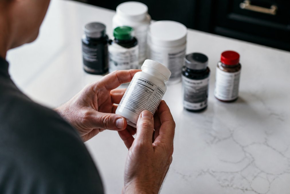 Close-up of a person's hands holding a supplement bottle, carefully reading the Supplement Facts label, with other supplement bottles arranged on a clean white countertop in soft natural light