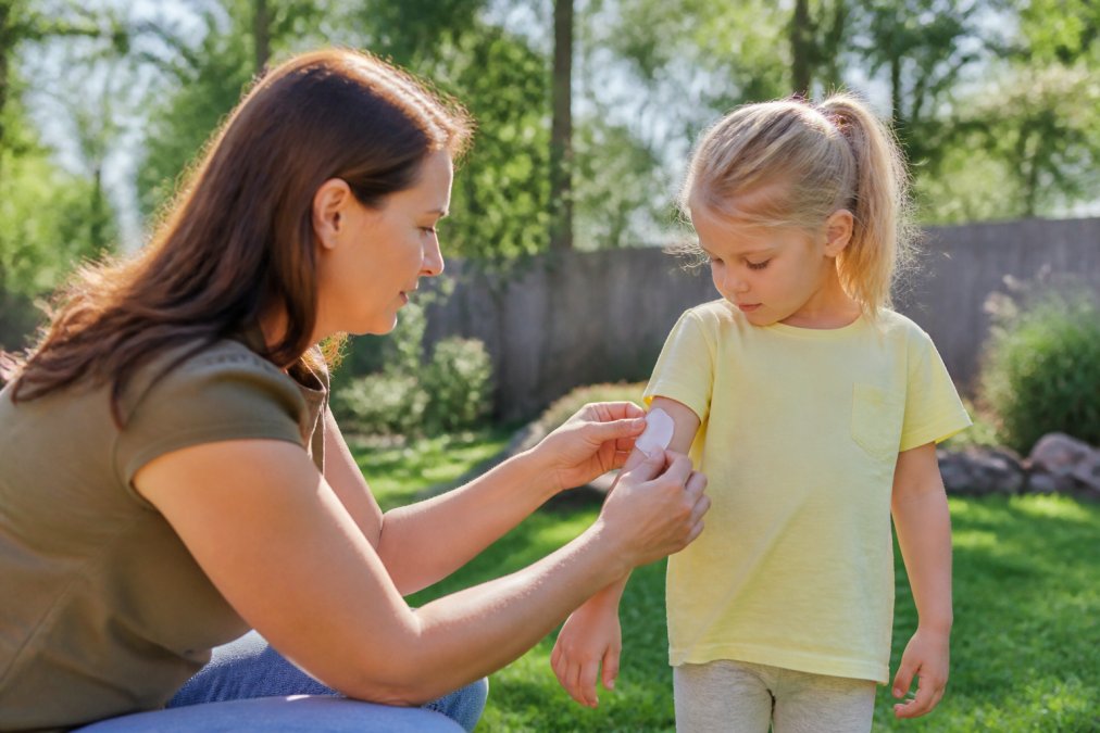 Parent carefully applying insect repellent patch to child's clothing sleeve while standing in a sunny backyard garden with green lawn visible