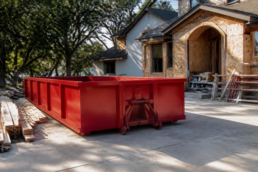 A red roll-off dumpster parked on a concrete residential driveway with a partially renovated house visible in the background, lumber and debris stacked nearby under afternoon sunlight