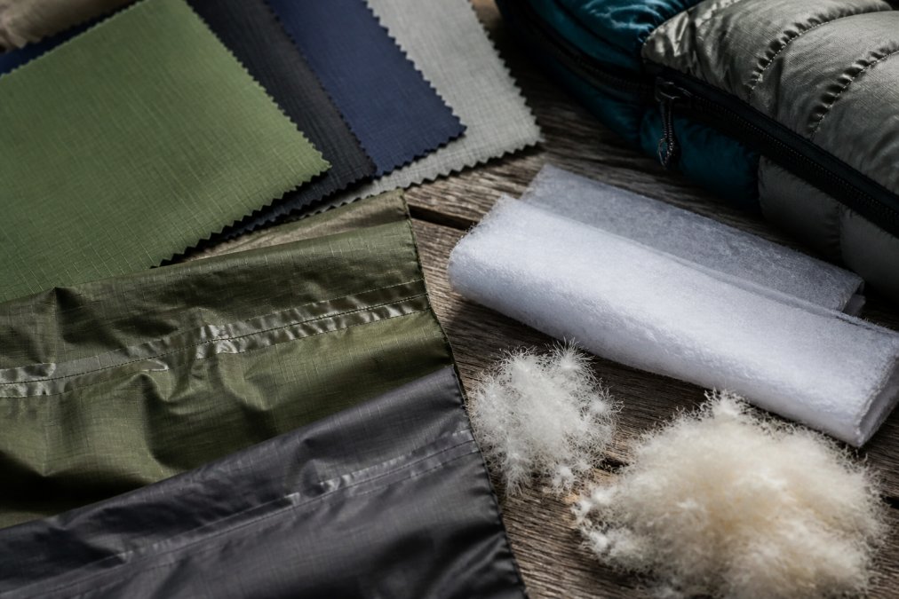 Close-up of various camping gear materials including ripstop nylon samples in different deniers, seam-sealed fabric sections, synthetic insulation batting, and down fill clusters arranged on a weathered outdoor surface with natural daylight