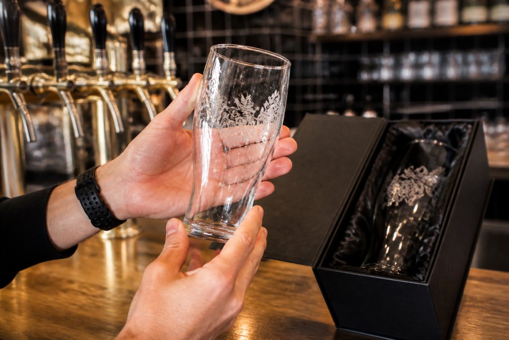 Close-up of hands holding a custom-etched beer glass under bright taproom lighting, showing the etched logo detail and condensation on the glass surface