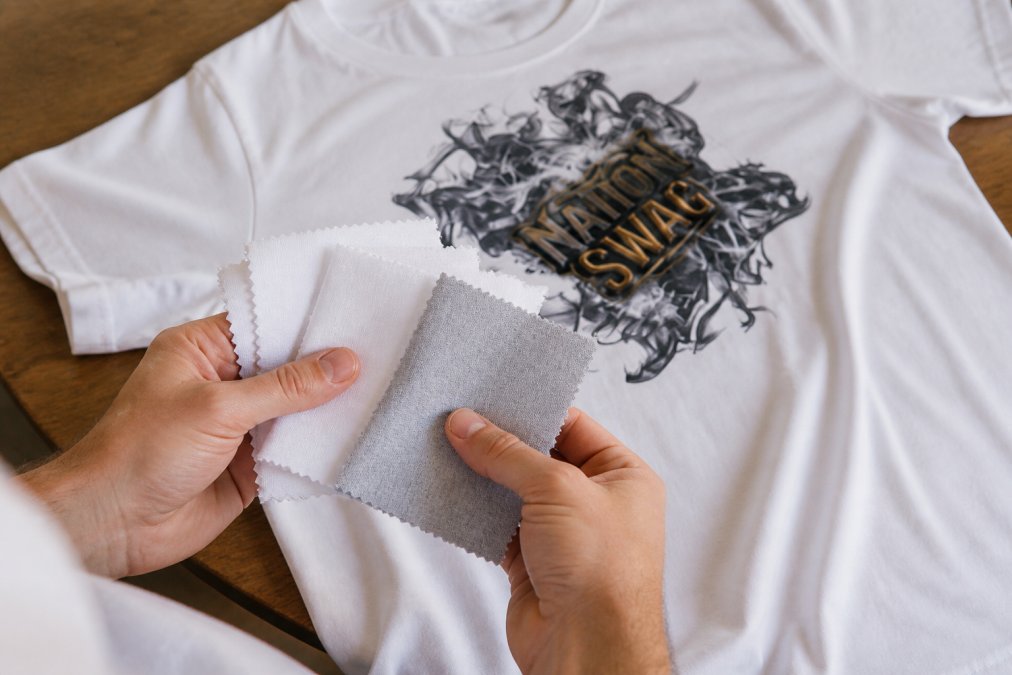 Close-up of hands holding and examining different fabric swatches showing texture differences between ring-spun cotton, combed cotton, and cotton-polyester blend materials in natural daylight