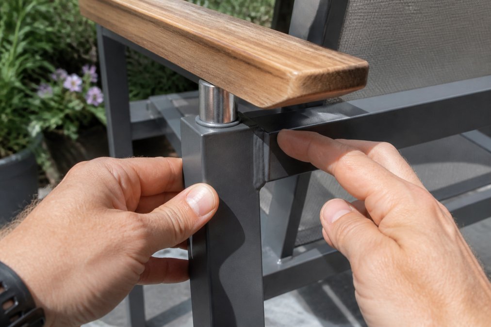 Close-up of hands inspecting a welded aluminum joint on a patio chair frame, with a teak wood armrest and stainless steel hardware visible in soft outdoor daylight