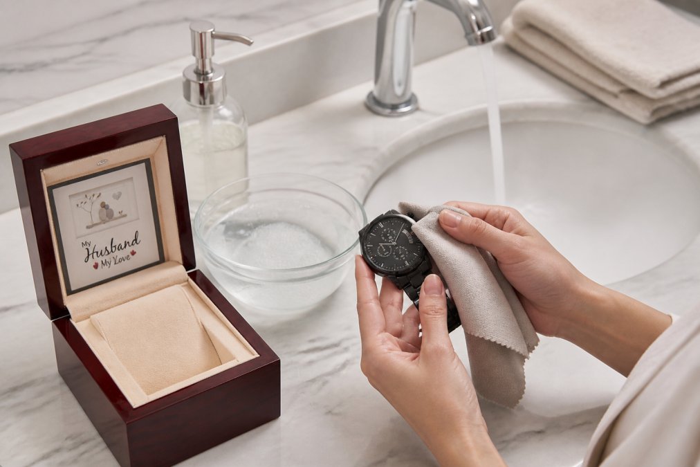 Person gently cleaning a personalized necklace with a soft microfiber cloth and mild soap solution at a bathroom sink, with warm water running gently in background