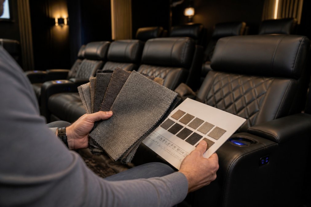 Person's hands holding multiple fabric swatches next to a durability rating card and cleaning code guide, seated in a modern home theater room with reclined seating visible in soft ambient lighting