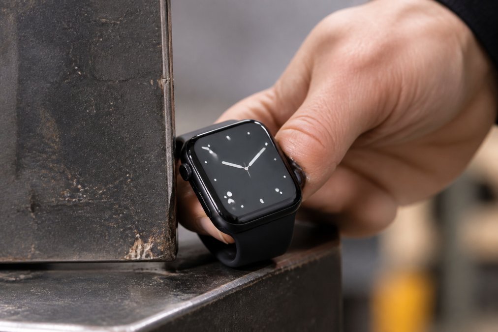 Close-up of a wearable device's protective corner reinforcement being tested against a rugged metal surface, showing hands positioning a smartwatch to demonstrate durability features during impact testing