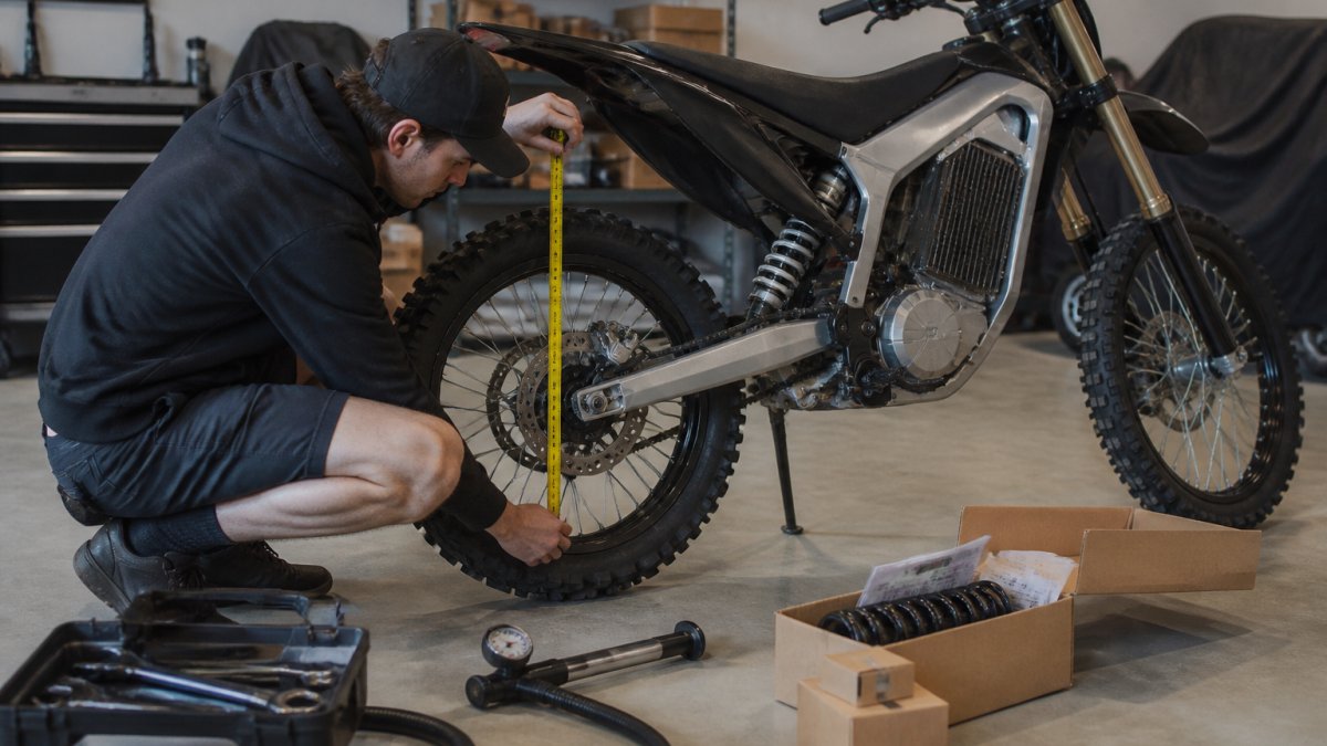 A rider crouching beside an electric dirt bike in a garage, measuring rear suspension sag with a tape measure against the rear axle, tools and a shock pump on the floor nearby