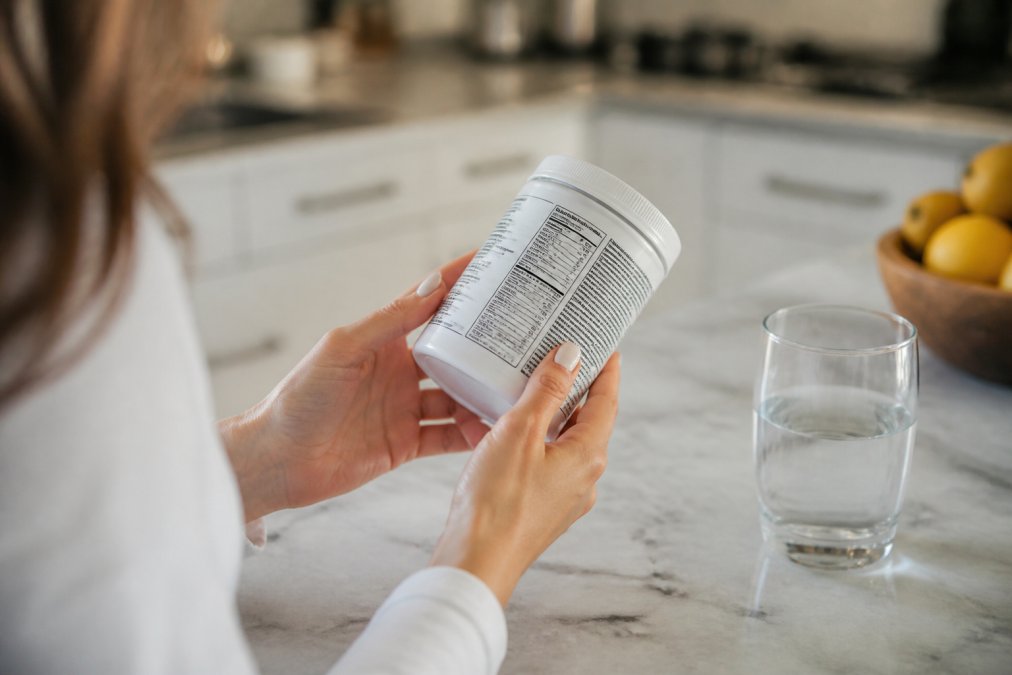 Close-up of a person's hands holding a supplement container and reading the ingredient label under bright natural kitchen light, with a glass of water nearby