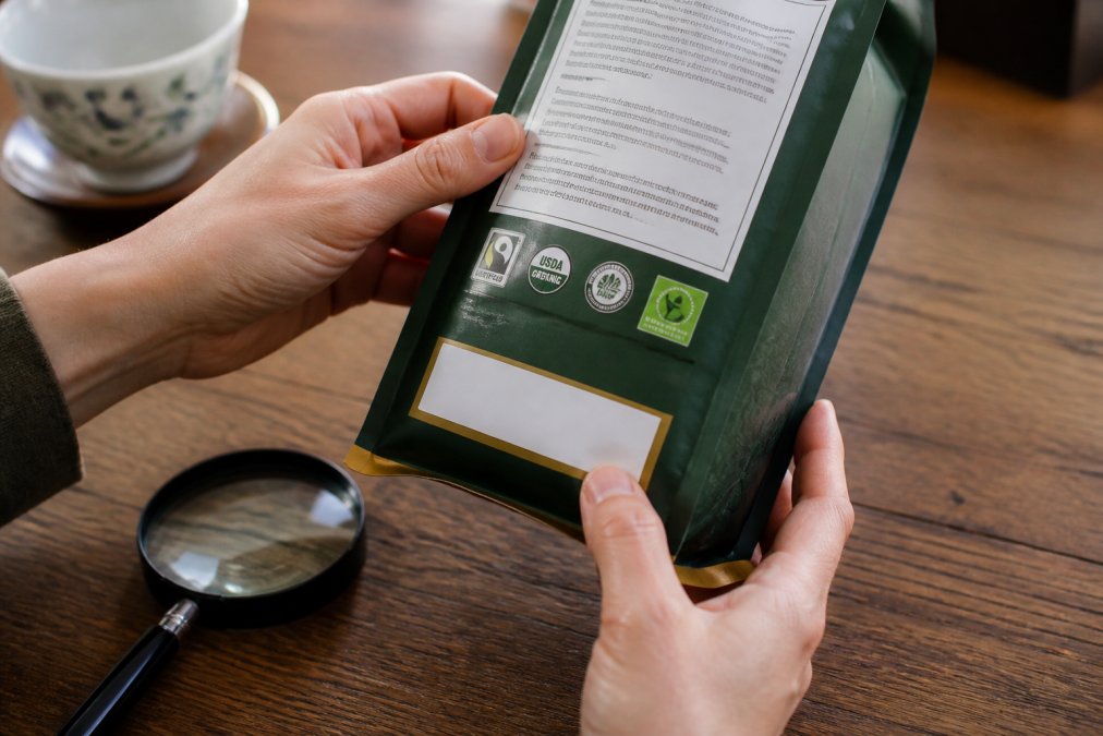 Close-up of hands holding a tea package, examining the certification labels and text on the back, with a magnifying glass nearby on a wooden table