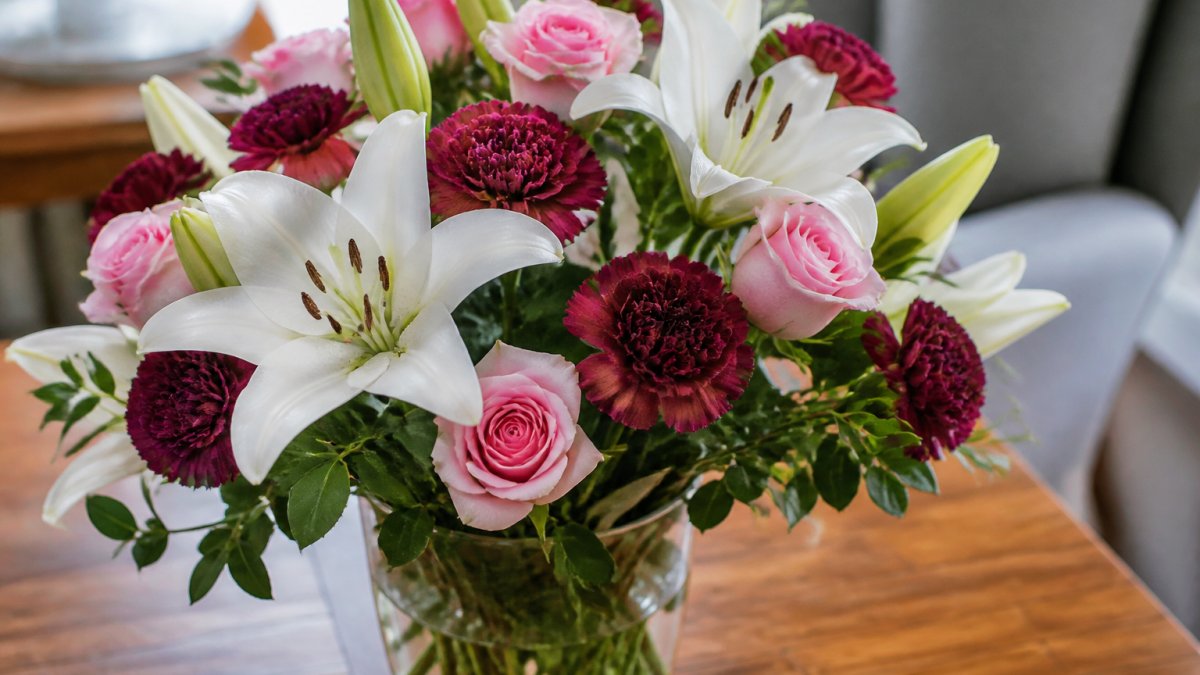 Close-up of white lilies with prominent stamens, pink roses, and burgundy carnations arranged in a clear glass vase with green foliage, set on a wooden table with soft natural lighting