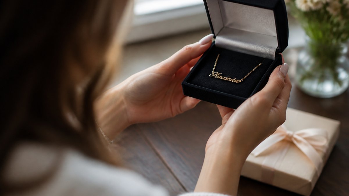 Close-up of a woman's hands opening a small jewelry box containing a delicate gold name necklace, soft natural window light, warm and intimate mood