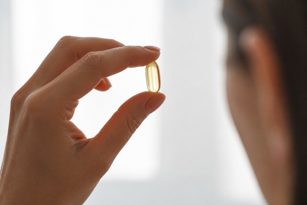 Person holding a fish oil softgel capsule up to bright light, examining its color and clarity to assess freshness and quality, with a clean white background