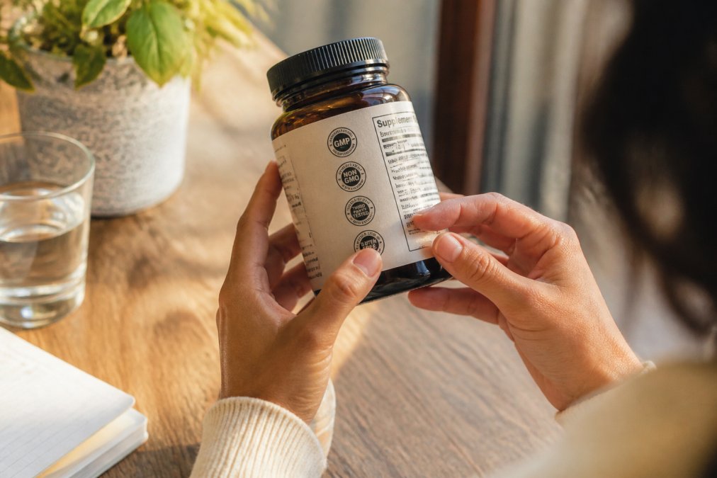 Close-up of hands holding a supplement bottle, examining certification seals and labels in bright natural light on a wooden surface